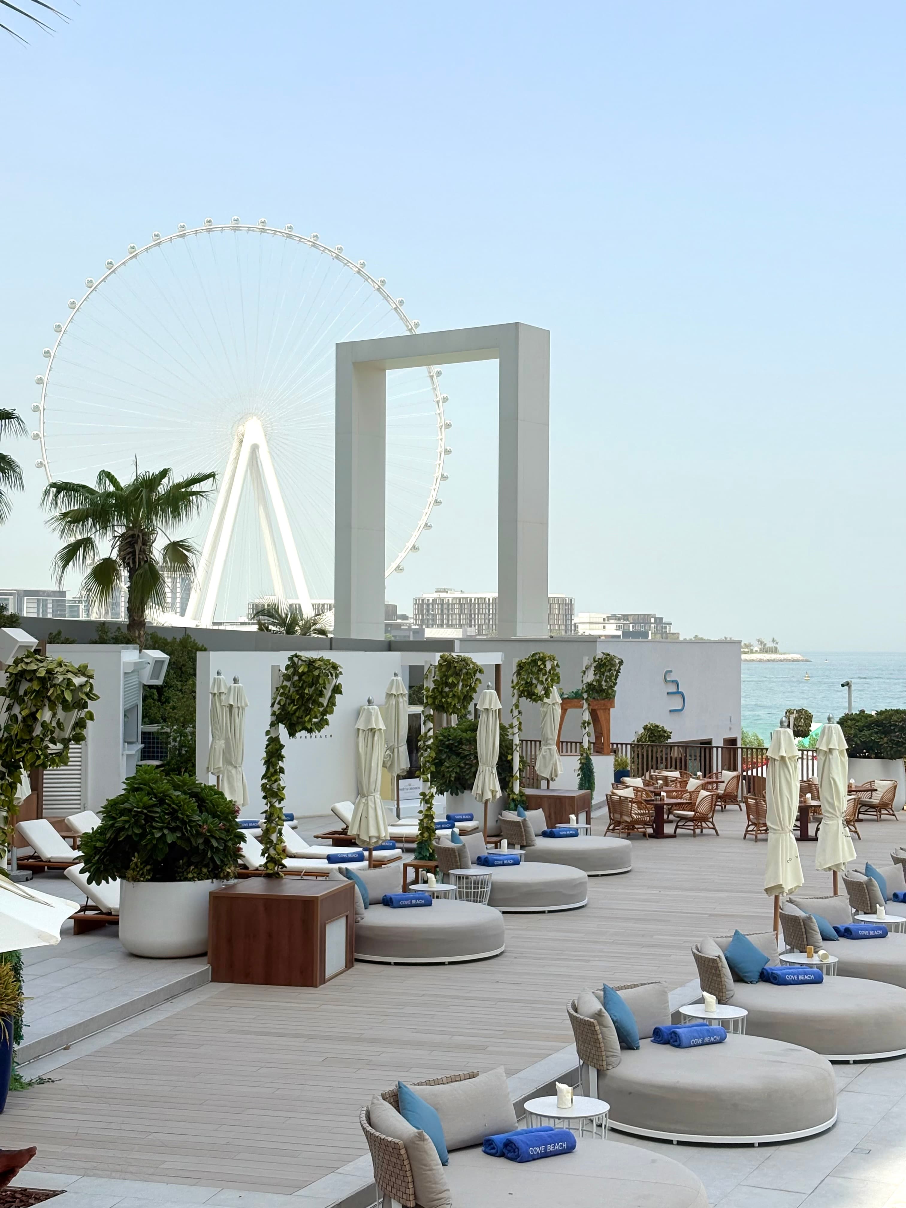 Beach club terrace featuring circular daybeds, white umbrellas, and the Ain Dubai Ferris wheel.