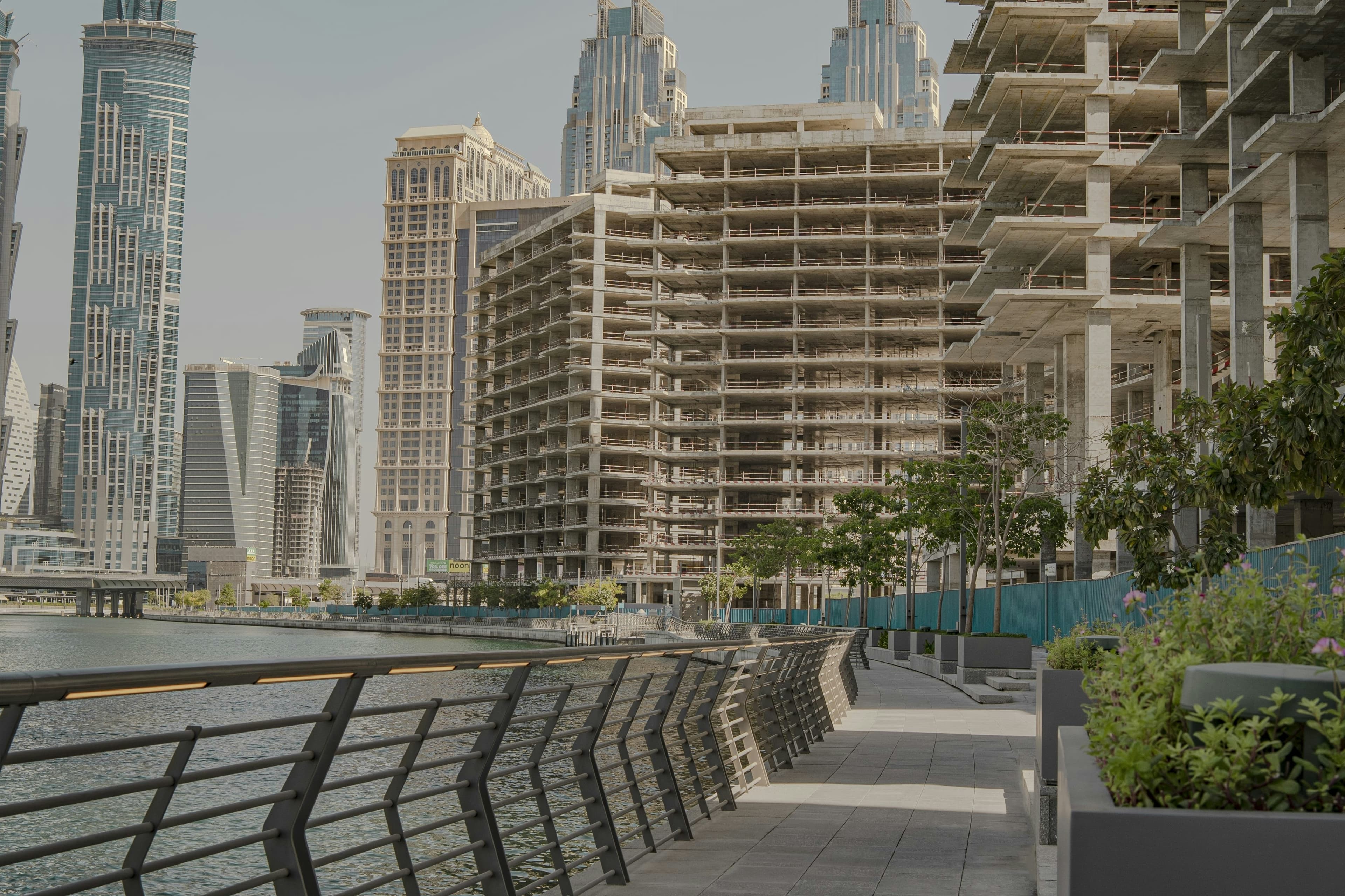 Paved waterfront walkway with metal railings beside concrete building skeletons and finished modern city skyscrapers.