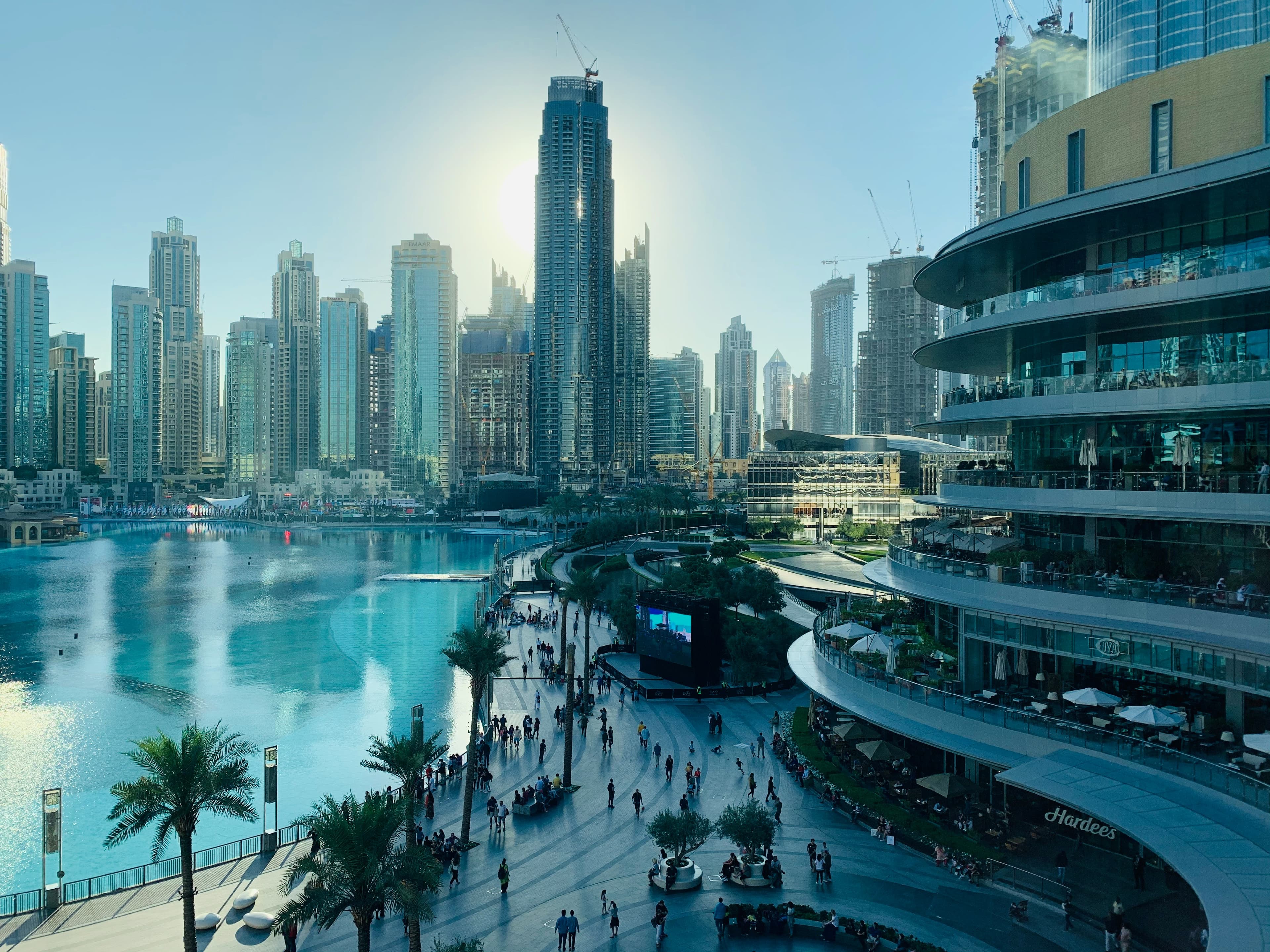 Sunlit skyscrapers overlook a turquoise lake and a bustling pedestrian promenade with palm trees.